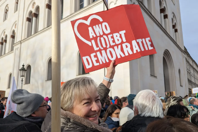 Eine ältere Frau hält bei einer Demonstration draußen ein rotes Schild hoch, auf dem in weißer Schrift steht: 